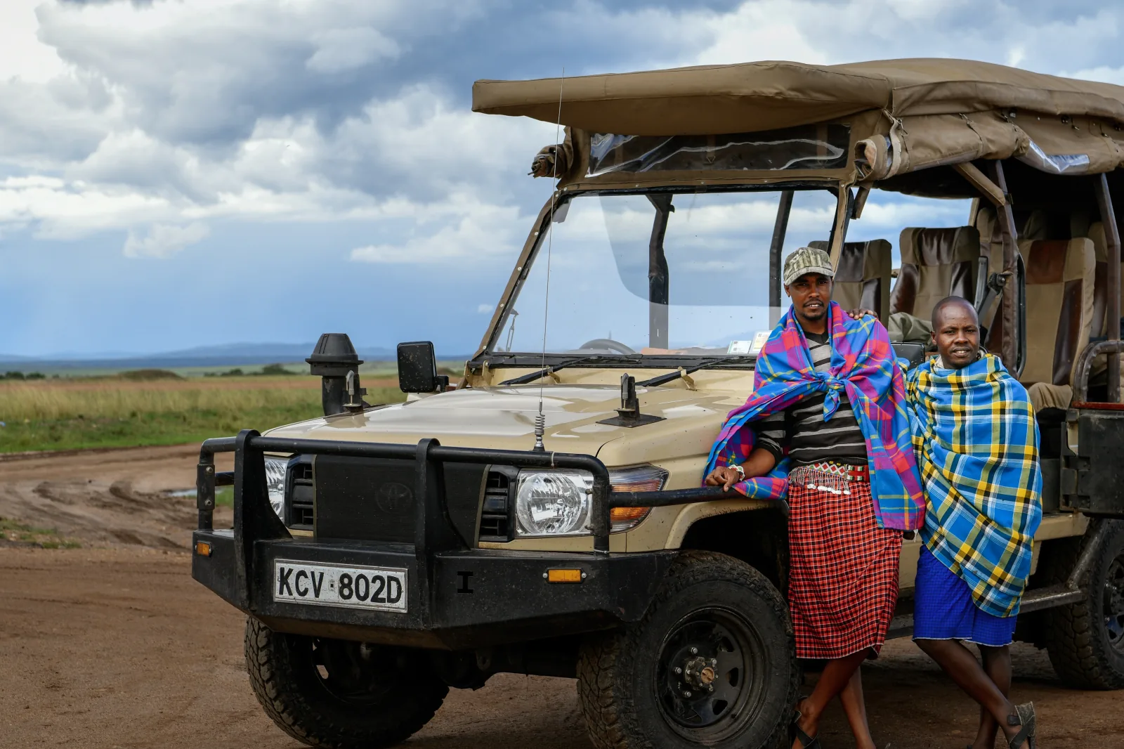 Två guider i rutiga masaifiltar lutar sig mot en öppen safaribil på en grusväg i Masai Mara