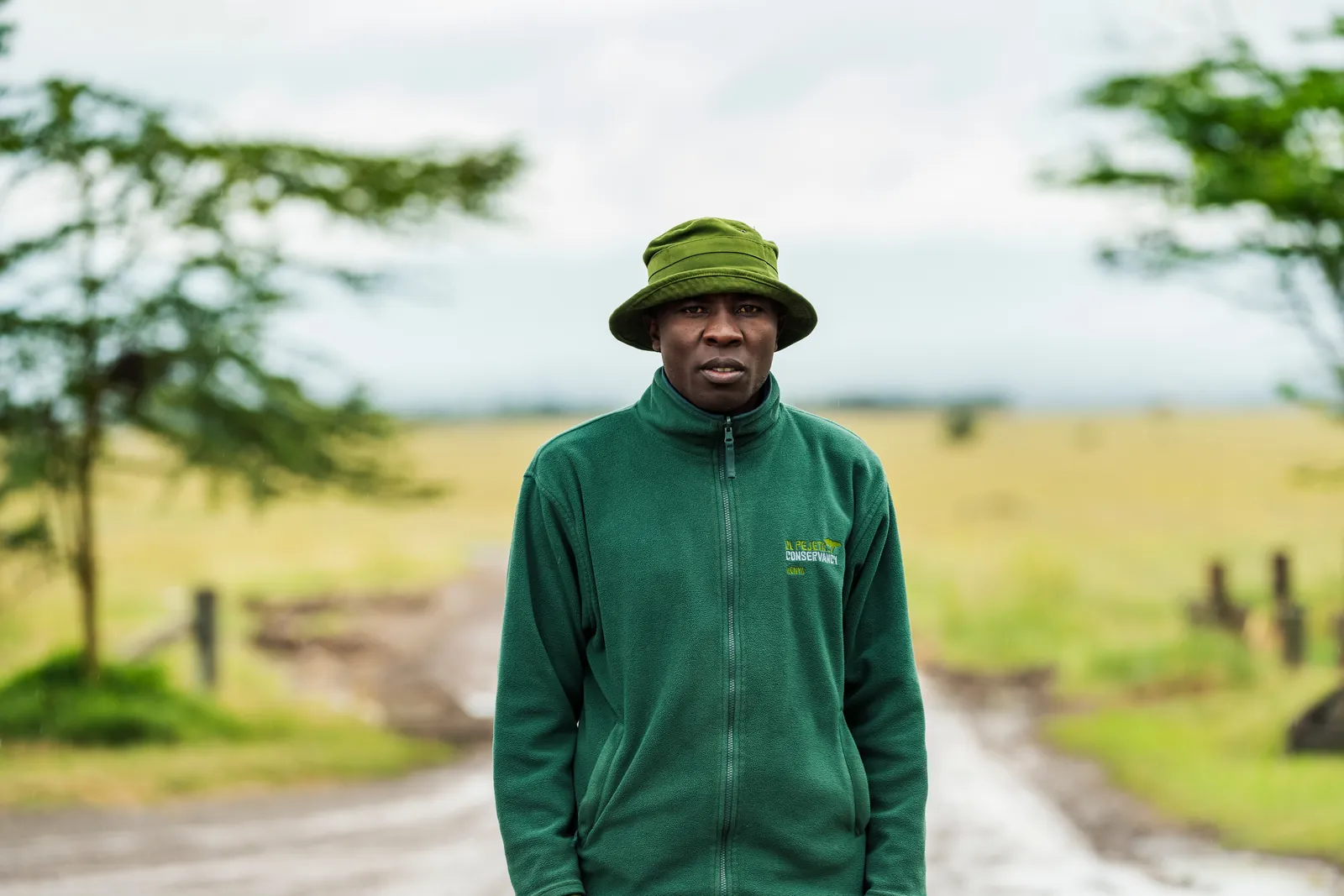 En man i grön fleecetröja och grön hatt står på en lerig väg mellan akacior och högt gräs. På tröjan står Ol Pejeta Conservancy.
